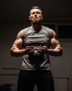 Muscular bearded man in a gym, wearing a gray sleeveless shirt, gripping a kettlebell with both hands, exuding strength, discipline, and determination.