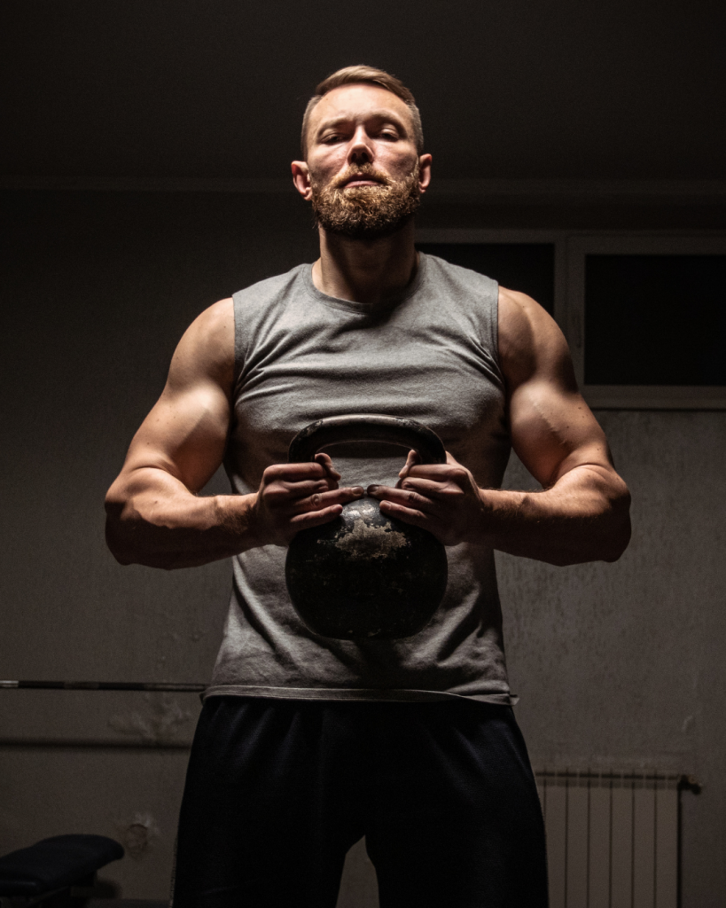 Muscular bearded man in a gym, wearing a gray sleeveless shirt, gripping a kettlebell with both hands, exuding strength, discipline, and determination.